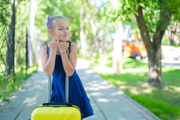 Een tevreden meisje in een blauwe jurk staat alleen op straat met een gele koffer en wacht op haar ouders. Een kind gaat op zomervakantie.. — Stockfoto