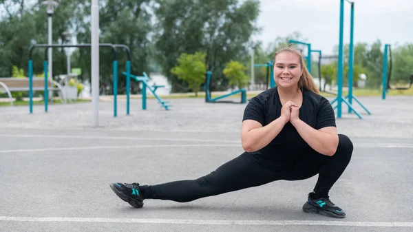 Fettleibige junge Kaukasierin beim Seitensprung. Dicke schöne lächelnde Mädchen in einem schwarzen Trainingsanzug ist in Fitness im Freien beschäftigt. Aufwärmen vor dem Training. — Stockfoto