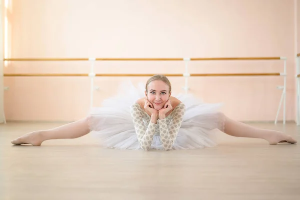 Bela bailarina em corpo e tutu branco está treinando em uma aula de dança. Jovem dançarina flexível posando em sapatos pontiagudos sentados no chão. — Fotografia de Stock