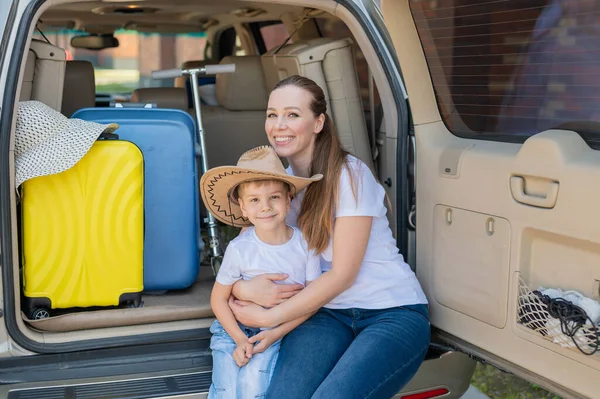 Europäerin sitzt mit ihrem Sohn im Kofferraum eines Geländewagens. Schöne glückliche Mutter liebevoll umarmt Sohn. Gelbes Gepäck für den Sommerurlaub mit dem Auto. Urlaub mit der ganzen Familie. — Stockfoto