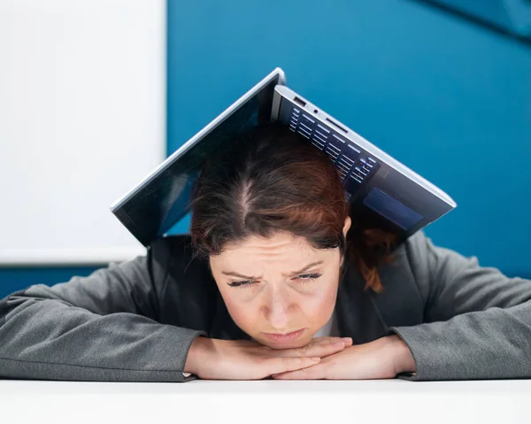 Problemer på jobben. En utslitt kvinne brettet laptopen sin over hodet ved pulten sin. Forretningskvinne i frustrasjon begravd under datamaskinen. Reduksjon og arbeidsledighet. – stockfoto