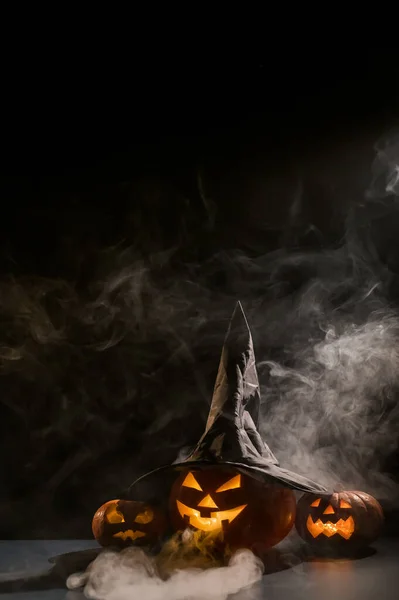 Vertikální Halloweenská karta. Čarodějnický klobouk na dýni s vyřezávanými úšklebky na černém pozadí v mlze. Jack-o-lucerna svítí zevnitř a vypouští kouř. — Stock fotografie