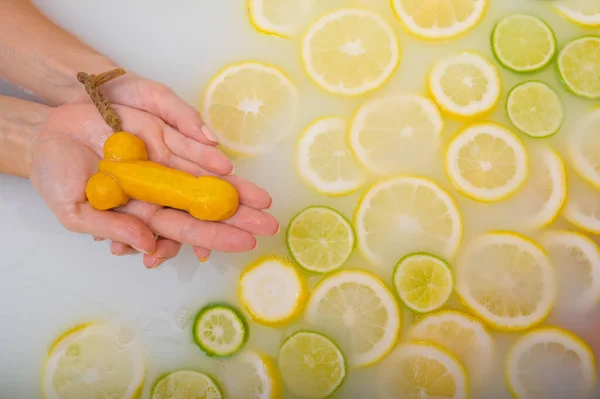 Fille prend un bain de lait avec des citrons et des citrons verts. Spa aux agrumes. Soins du corps. Blanchiment de la peau. Une femme se lave les mains avec du savon sous la forme d'un pénis masculin. Humour . — Photo