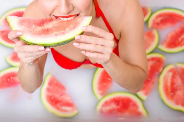 Porträt einer Frau im roten Badeanzug, die eine Wassermelone isst und grinst. Rotschopf Mädchen mit rotem Lippenstift nimmt ein Bad mit Milch und Früchten, um ihre Haut zu verjüngen. Körperpflege. — Stockfoto