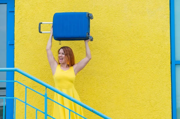 穿着黄色衣服的快乐女人头顶着一个蓝色的手提箱，靠着一面黄色的墙。白人女孩笑着，快乐地等待着旅行. — 图库照片