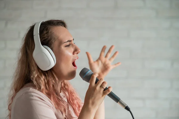 Közelkép egy göndör hajú kaukázusi nőről, aki mikrofonba énekel. Gyönyörű érzelmi lány fehér fülhallgatóval énekel egy dalt otthon karaoke és aktívan gesztusok ellen téglafal. — Stock Fotó