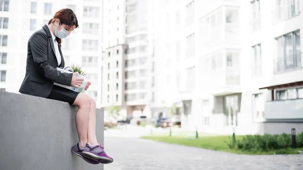 Mujer deprimida en máscara médica se sienta en el parapeto al aire libre con una caja de artículos personales desde el escritorio. Una chica desempleada sola en la calle. Quiebra de una empresa por cuarentena en coronavirus. —  Fotos de Stock