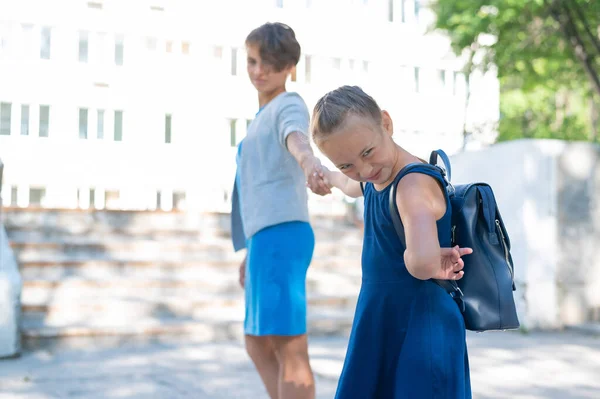 Uma menina infeliz com uma mochila puxa a mãe pela mão não quer ir à escola. Uma mulher leva uma filha que resiste à escola. O conflito de gerações. — Fotografia de Stock