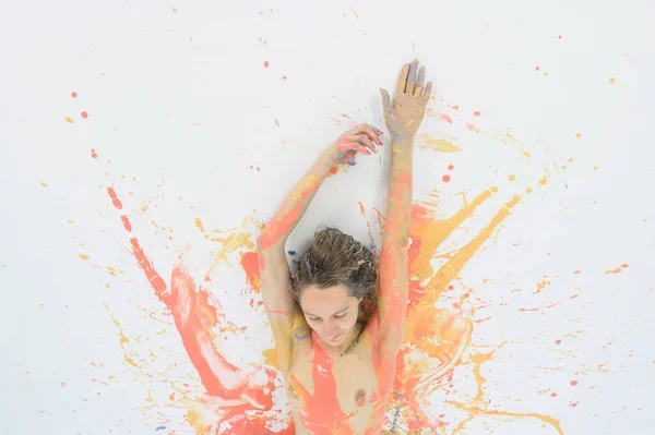 Vista superior de una mujer desnuda en un spray de pintura. Una chica sin ropa yace en un suelo blanco en manchas y manchas de colores. Arte corporal extraño. . —  Fotos de Stock