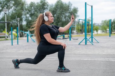 Kulaklık takan güzel şişman bir kız spor sahasında spor yapıyor ve selfie çekiyor. Genç bir kadın saldırır ve sıcak bir yaz gününde açık havada akıllı bir telefonla fotoğraf çekilir..