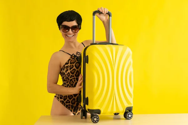 튼튼 한 표범 수영복을 입고 선글라스를 쓴 여자가 노란 배경에 여행 가방을 들고 있다. 단 키니를 입고 미소짓고 있는 갈색 머리 새가 가방을 싸서 바다로 여행 할 준비가 되어 있다. — 스톡 사진
