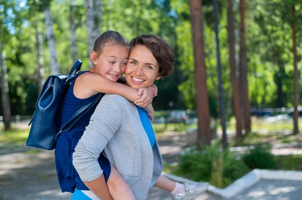 En smuk hvid kvinde står udendørs og holder datter på ryggen. En munter smilende pige sidder på sin mor tilbage, mens du går i parken. Venlig kærlig familie. - Stock-foto