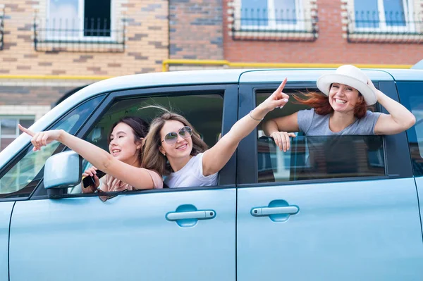 Drei fröhliche kaukasische Freundinnen blicken aus dem Autofenster. Aufgeregte Frauen gehen auf Roadtrip. Die brünette Blondine und der Rotschopf machen Urlaub. — Stockfoto