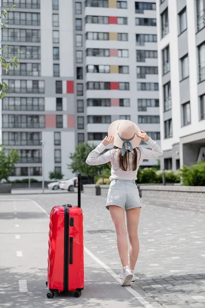 Ein Mädchen in kurzen Hosen und Hut steht mit einem roten Koffer draußen. Eine junge Frau läuft an einem sonnigen Sommertag mit Gepäck eine leere Straße entlang. Alleinreisende mit einer großen Tasche. — Stockfoto