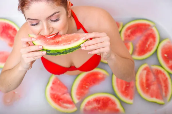 Porträt einer Frau im roten Badeanzug, die eine Wassermelone isst und grinst. Rotschopf Mädchen mit rotem Lippenstift nimmt ein Bad mit Milch und Früchten, um ihre Haut zu verjüngen. Körperpflege. — Stockfoto