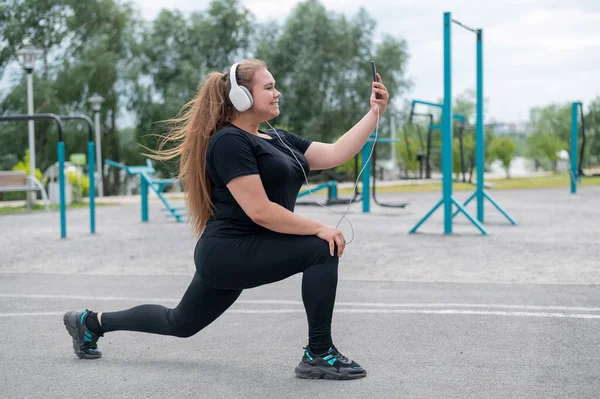 Ein hübsches, dickes Mädchen mit Kopfhörern übt sich auf dem Sportplatz in Fitness und macht ein Selfie. Junge Frau stürzt und wird an einem warmen Sommertag im Freien mit dem Smartphone fotografiert. — Stockfoto