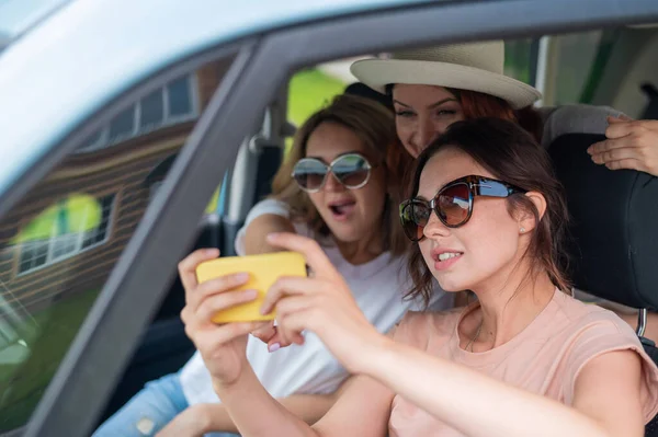 Três namoradas vão em uma viagem e tiram uma selfie em um smartphone. A mulher está sentada no carro e tirando fotos com seu celular. Viagem de férias . — Fotografia de Stock