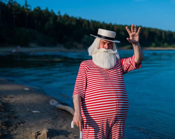 Un anziano uomo dai capelli grigi in costume da bagno classico e cappello si erge sulla spiaggia con un ombrellone e saluta con la mano. In pensione con la barba in vacanza. — Foto Stock