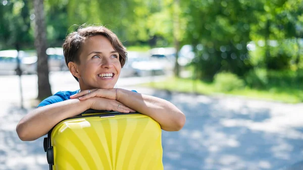 Gros plan portrait d'une belle jeune femme blanche aux cheveux courts avec une valise jaune à l'extérieur. La fille au sourire parfait se pencha la tête contre un grand sac à bagages. — Photo