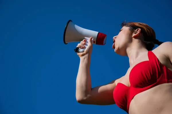 Белая женщина в красном бикини работает спасателем на пляже и кричит через мегафон о голубое небо. Девушка в купальнике стоит с громкоговорителем. Летний отдых. — стоковое фото