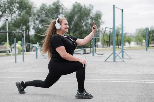 Ein hübsches, dickes Mädchen mit Kopfhörern übt sich auf dem Sportplatz in Fitness und macht ein Selfie. Junge Frau stürzt und wird an einem warmen Sommertag im Freien mit dem Smartphone fotografiert. — Stockfoto