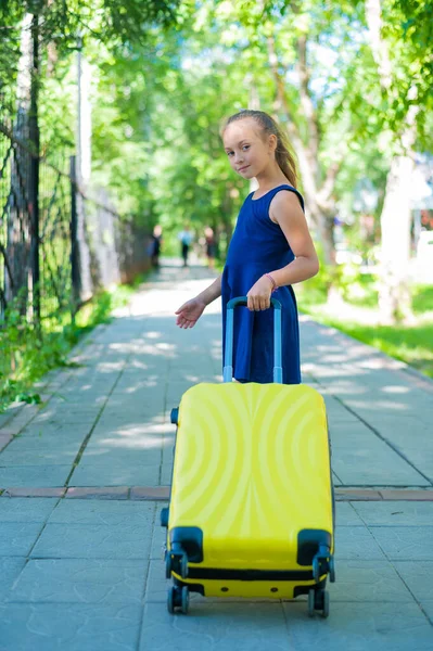 Een lachend klein meisje in een blauwe jurk loopt langs de straat met een gele koffer. Een kind gaat op zomervakantie.. — Stockfoto
