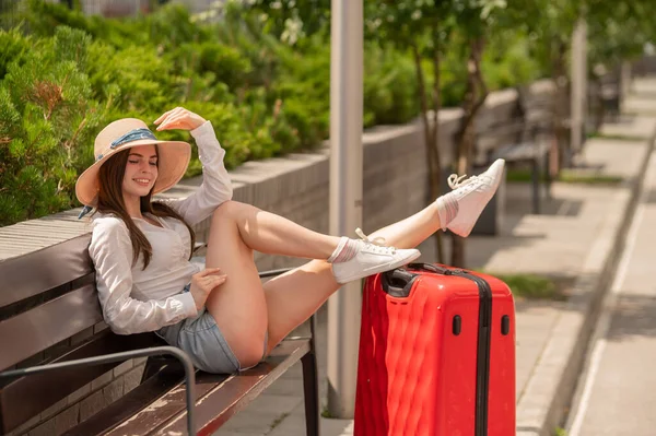 Mooie brunette in korte broek en een hoed zit op een bank gestrekte benen op een grote rode koffer. Blanke vrouw vouwde haar voeten op een tas buiten. Meisje geniet van een reis in het park op een zonnige. — Stockfoto