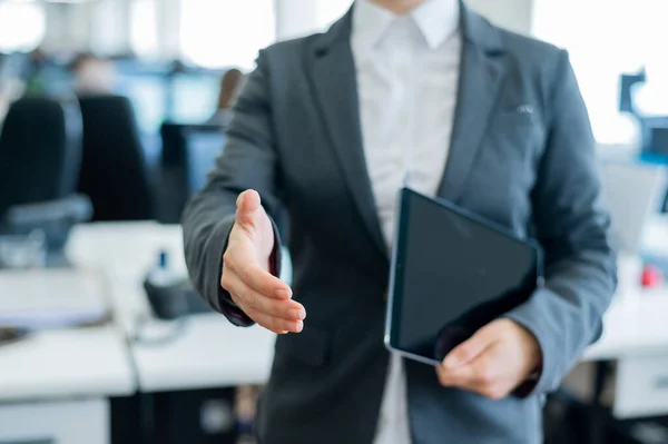 Business woman in a jacket and white shirt holding out a hand for a greeting. A faceless business woman holds a digital tablet and shakes her hand as an agreement on a successful transaction.