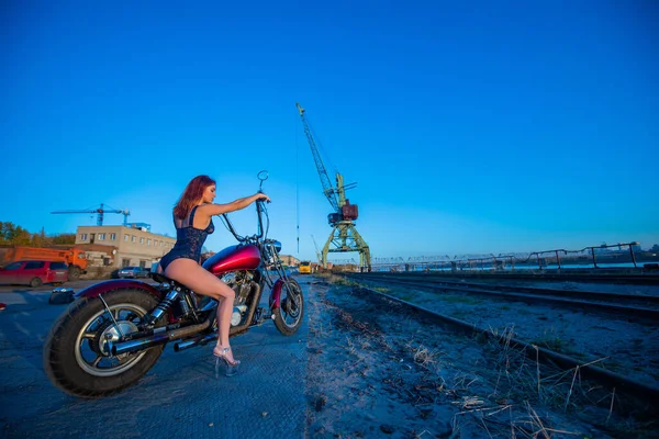 Sexy jovem mulher ruiva em roupa interior erótica senta-se em uma motocicleta vermelha. Menina sedutora dirige uma bicicleta em um fundo industrial . — Fotografia de Stock