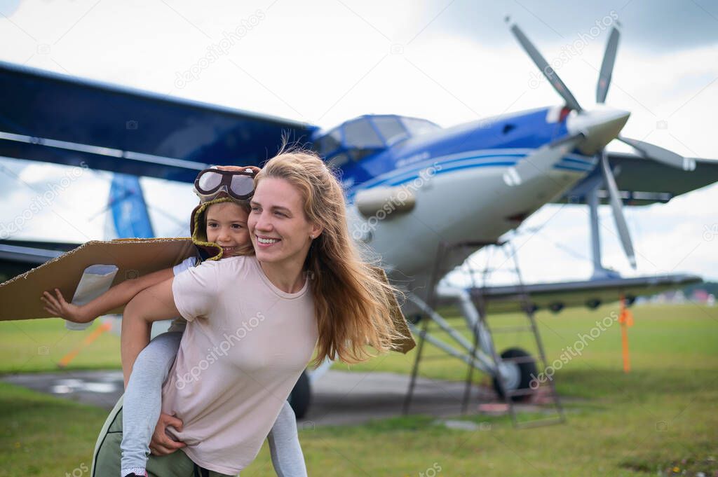 Beautiful caucasian woman is holding her little daughter at the airfield. A small girl in a pilots costume sits on her mothers back against the background of the plane.