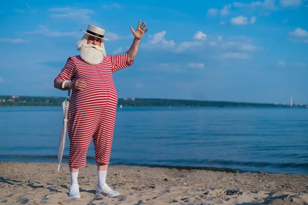 Un anziano uomo dai capelli grigi in costume da bagno classico e cappello si erge sulla spiaggia con un ombrellone e saluta con la mano. In pensione con la barba in vacanza. — Foto Stock