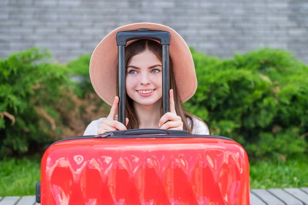 Joven hermosa mujer caucásica en un sombrero se ve fuera de una maleta roja con un mango. La chica feliz va de viaje con el equipaje. Concepto vacaciones de verano. — Foto de Stock