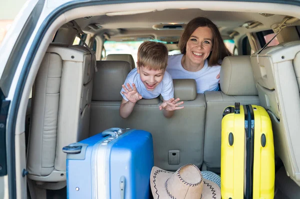 Glückliche Europäerin mit ihrem kleinen Sohn blickt auf den Kofferraum eines Autos mit Koffern. Sommerurlaub für die Familie. Mit einem Kind im Auto unterwegs. Ein Junge mit seiner Mutter auf dem Rücksitz eines Jeeps. — Stockfoto