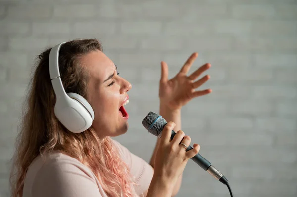 Närbild av en kaukasisk kvinna med lockigt hår som sjunger in i en mikrofon. Vacker känslomässig flicka i vita hörlurar sjunger en sång i hemmet karaoke och aktivt gester mot en tegelvägg. — Stockfoto