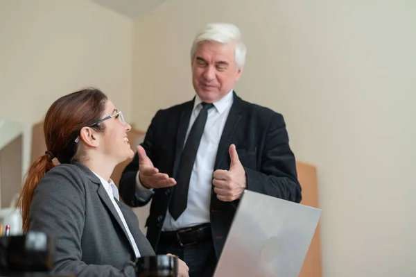 Ein reifer männlicher Chef lobt einen Untergebenen. Eine Frau im Anzug arbeitet an einem Laptop am Schreibtisch. Freundliche Kollegen plaudern im Büro. — Stockfoto