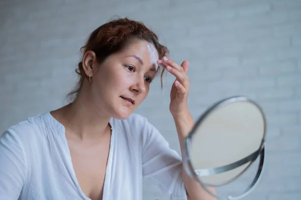 Retrato de una mujer caucásica con vitíligo utiliza protector solar. Una chica con una mancha de pigmento blanco en la frente se mira en el espejo y se unta con crema. — Foto de Stock