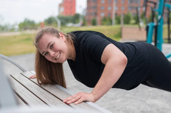 Schöne dicke junge Frau schiebt sich von der Bank nach oben. Ein pausbäckiges lächelndes Mädchen wärmt sich vor dem Joggen auf. Sport zur Gewichtsabnahme. — Stockfoto