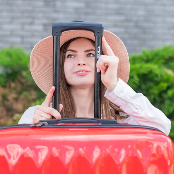 Junge schöne kaukasische Frau mit Hut schaut aus einem roten Koffer mit Griff. Glückliches Mädchen geht auf eine Reise mit Gepäck. Sommerferienkonzept. — Stockfoto