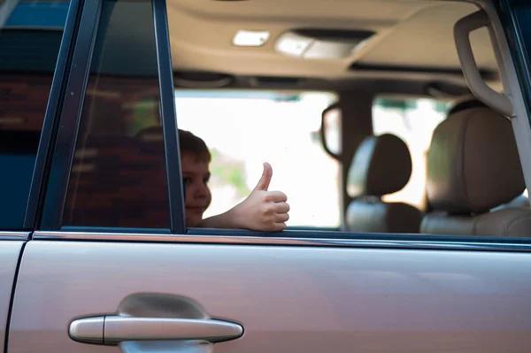 O garoto se senta no banco de trás de um carro prateado e mostra um polegar. Pré-escolar está pronto para uma viagem emocionante . — Fotografia de Stock
