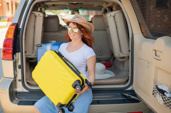 Glückliche rothaarige Frau mit Cowboyhut und Sonnenbrille auf einer Autofahrt durch das Land. Mädchen im Kofferraum mit gelbem Koffer, bereit für den Sommerurlaub. Unabhängiges Reisen. — Stockfoto