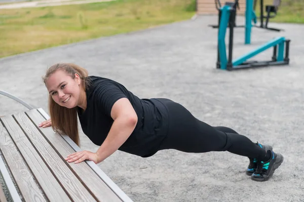 Tlustá mladá žena se zvedá z lavičky venku. Buclatá usměvavá dívka se před běháním zahřeje. Sport pro hubnutí. — Stock fotografie