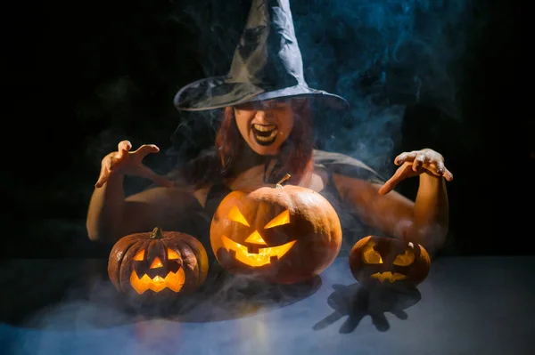 En ildevarslende heks i en hat fremmaner over en jack-o-lanterne. Traditionelle halloween tegn. Mystisk tåge kryber over græskar med udskårne forfærdelige ansigter. Kvinde i karneval kostume. - Stock-foto