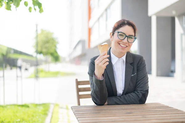 Die lächelnde Frau genießt es, an einem Holztisch im Freien eine Eistüte zu essen. Porträt eines Mädchens im Businessanzug für eine Pause in einem Straßencafé an einem heißen Sommertag. — Stockfoto