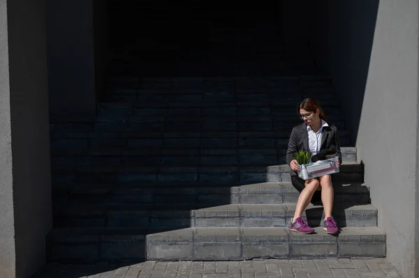 Žena v depresi, oblečená do lékařské masky, je propuštěna a sedí na schodech s krabicí osobních věcí. Dělnice v obleku a teniskách venku. Nezaměstnanost v hospodářské krizi — Stock fotografie