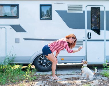 Güzel beyaz kadın karavanın yanında bir köpeği eğitiyor. Hayvanınla seyyar bir karavanla seyahat et. Kızıl saçlı kız Jack Russell Terrier ile oynuyor. Kadın karavan sürücüsü.
