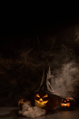 Dikey Cadılar Bayramı kartı. Sisteki siyah arka planda garip şekiller oyulmuş bir balkabağına cadı şapkası. Jack-o-fener içeriden parlar ve duman yayar.