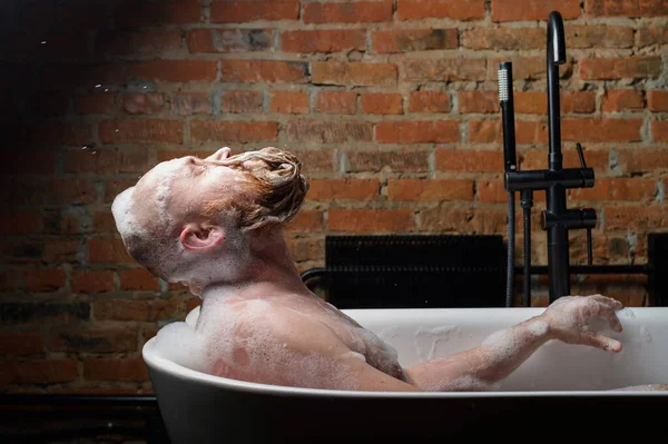 Close-up portrait of funny red-bearded bald man takes a bath with foam on the background of a brick wall. Cheerful naughty guy at spa treatments. A parody of glamorous girls. — Stock Photo, Image