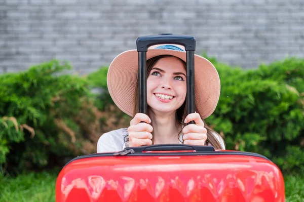 Jonge mooie blanke vrouw met een hoed kijkt uit een rode koffer met een handvat. Gelukkig meisje gaat op reis met bagage. Zomervakantie concept. — Stockfoto