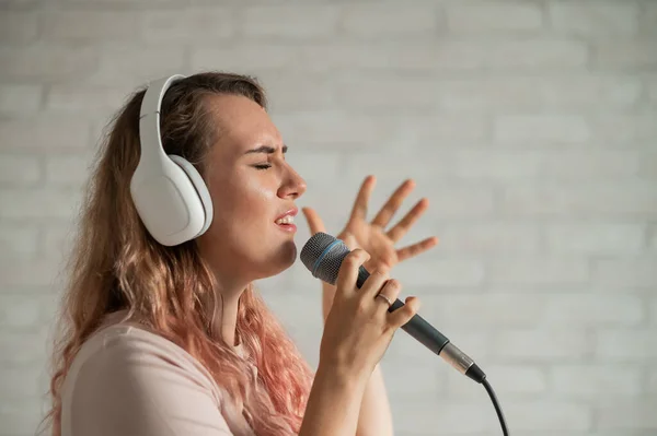 Großaufnahme Porträt einer kaukasischen Frau mit lockigem Haar, die in ein Mikrofon singt. Schöne emotionale Mädchen mit weißen Kopfhörern singt ein Lied in Heim-Karaoke und gestikuliert aktiv gegen eine Ziegelwand. — Stockfoto
