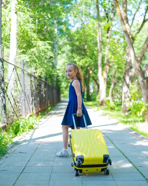 Een lachend klein meisje in een blauwe jurk loopt langs de straat met een gele koffer. Een kind gaat op zomervakantie.. — Stockfoto